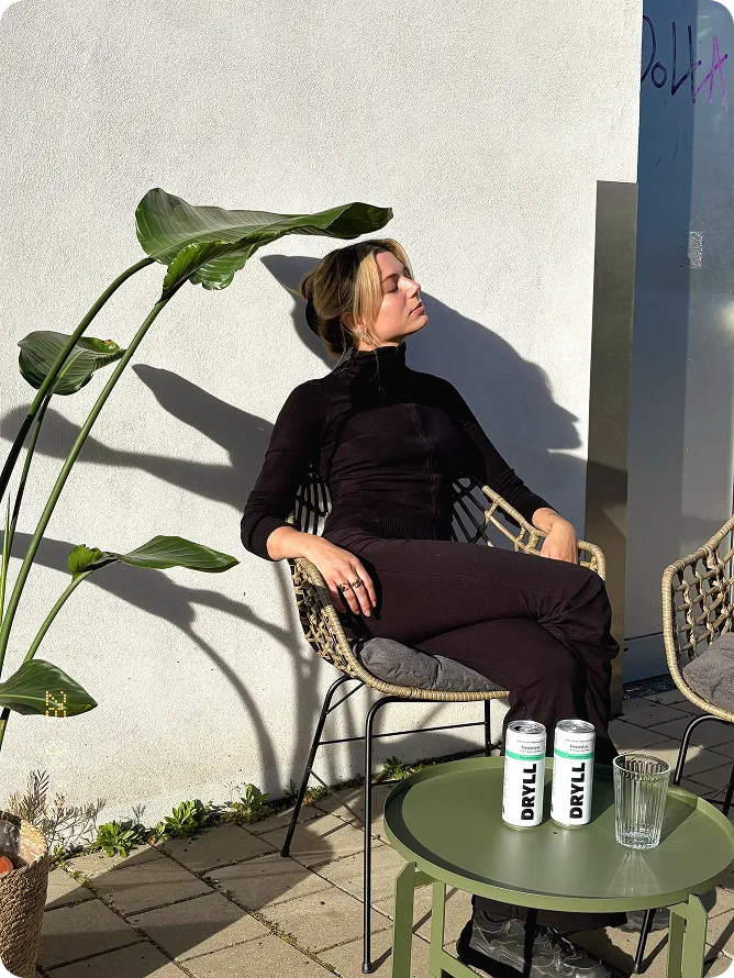 Woman in black sits on a woven chair, eyes closed, near green plant and table with drinks and glass.