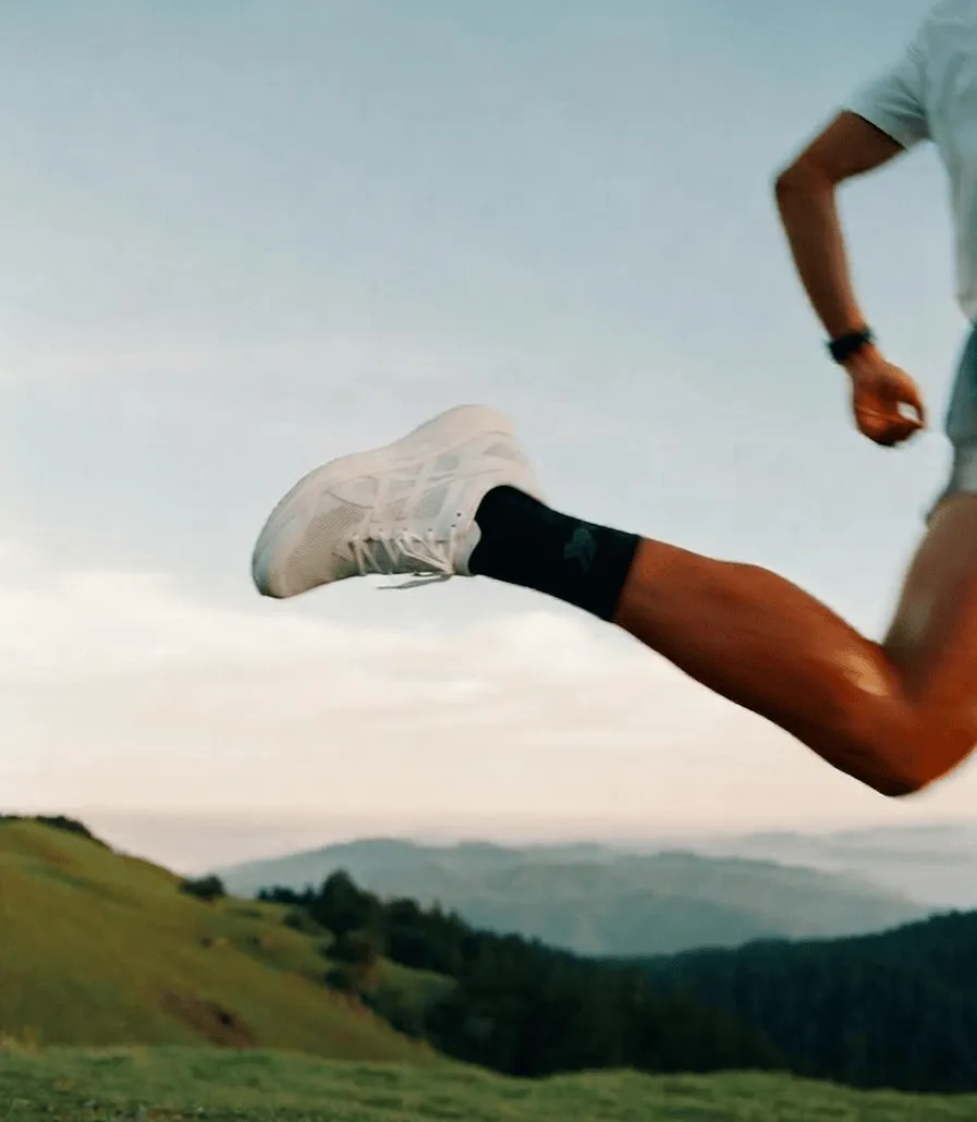 Person mid-jump wearing white sneakers and black socks over a grassy hillside with hills in background.