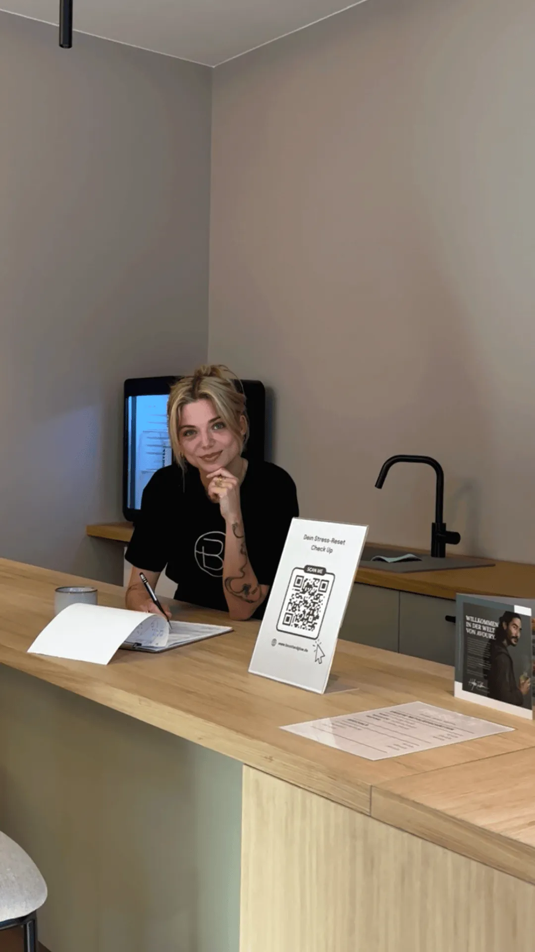Woman with blonde hair writing at a wooden counter with QR code sign and faucet in background.