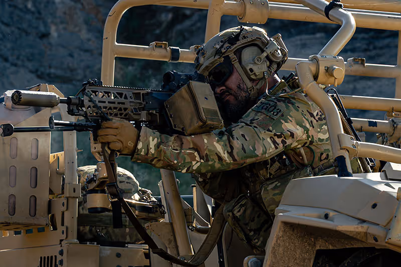 Soldier in camouflage aiming a rifle while seated in a tactical military vehicle. Brigantes