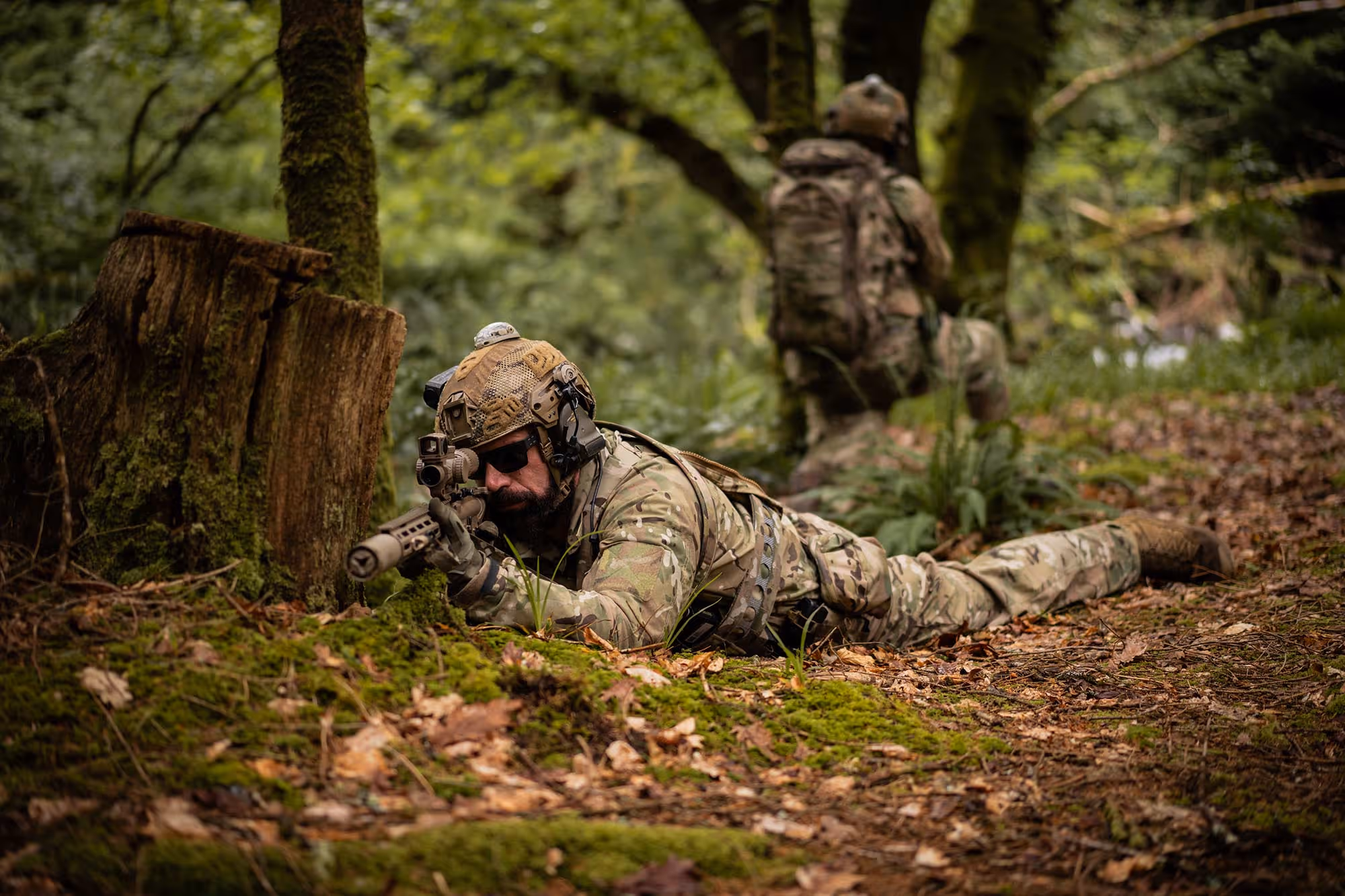 Soldier in camouflage gear lying on forest floor aiming a rifle, with another soldier kneeling in background. Brigantes