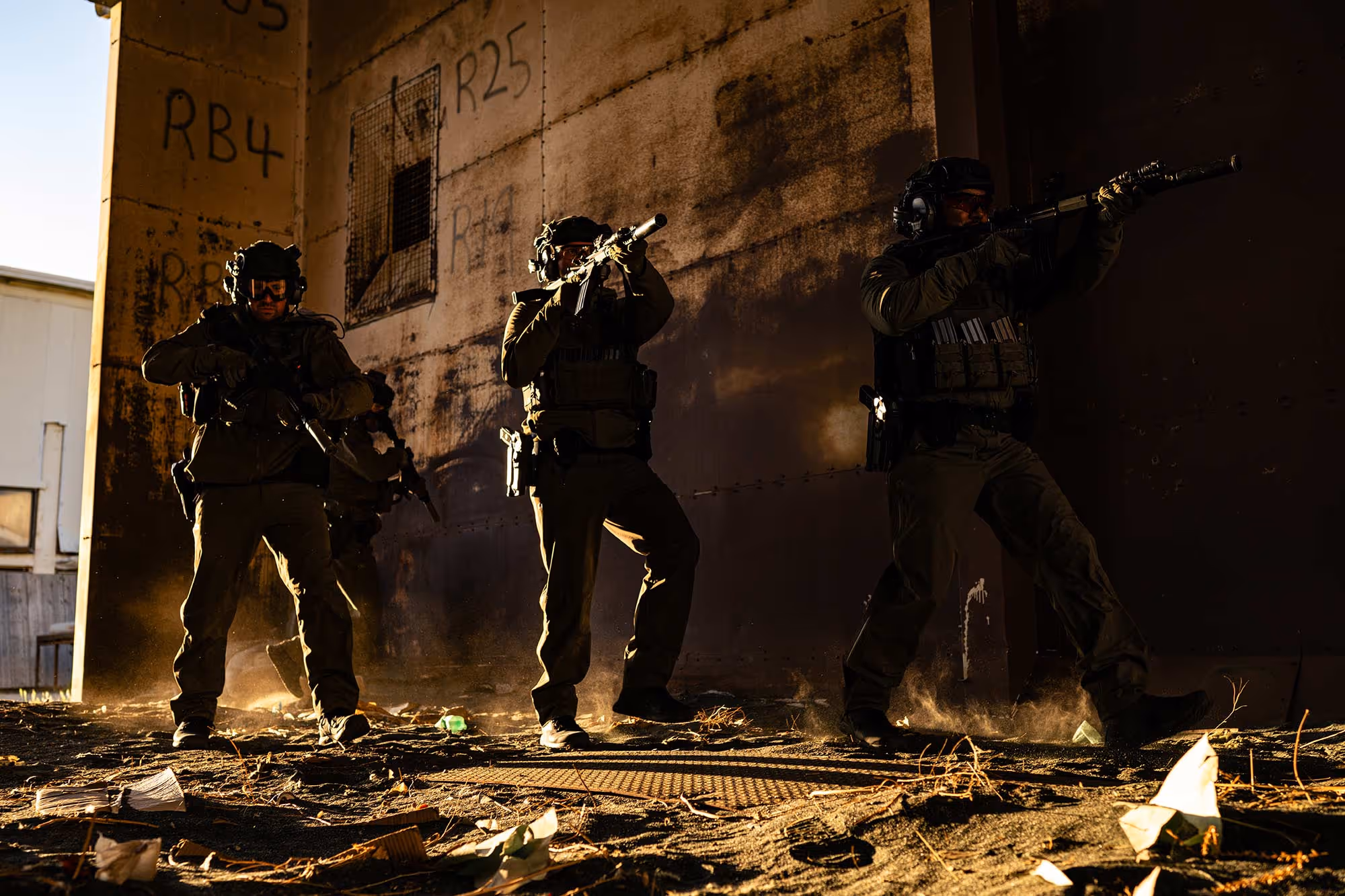 Four armed tactical officers in gear moving cautiously in a dusty, graffiti-marked urban area. Brigantes