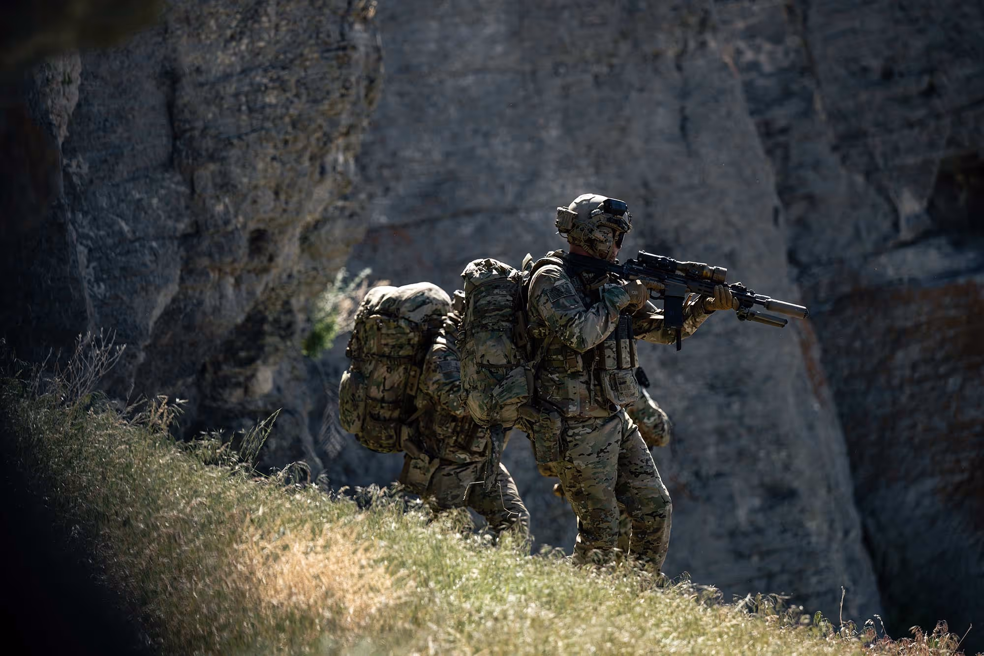 Two soldiers in camouflage gear walking on a grassy slope with rocky cliffs in the background. Brigantes