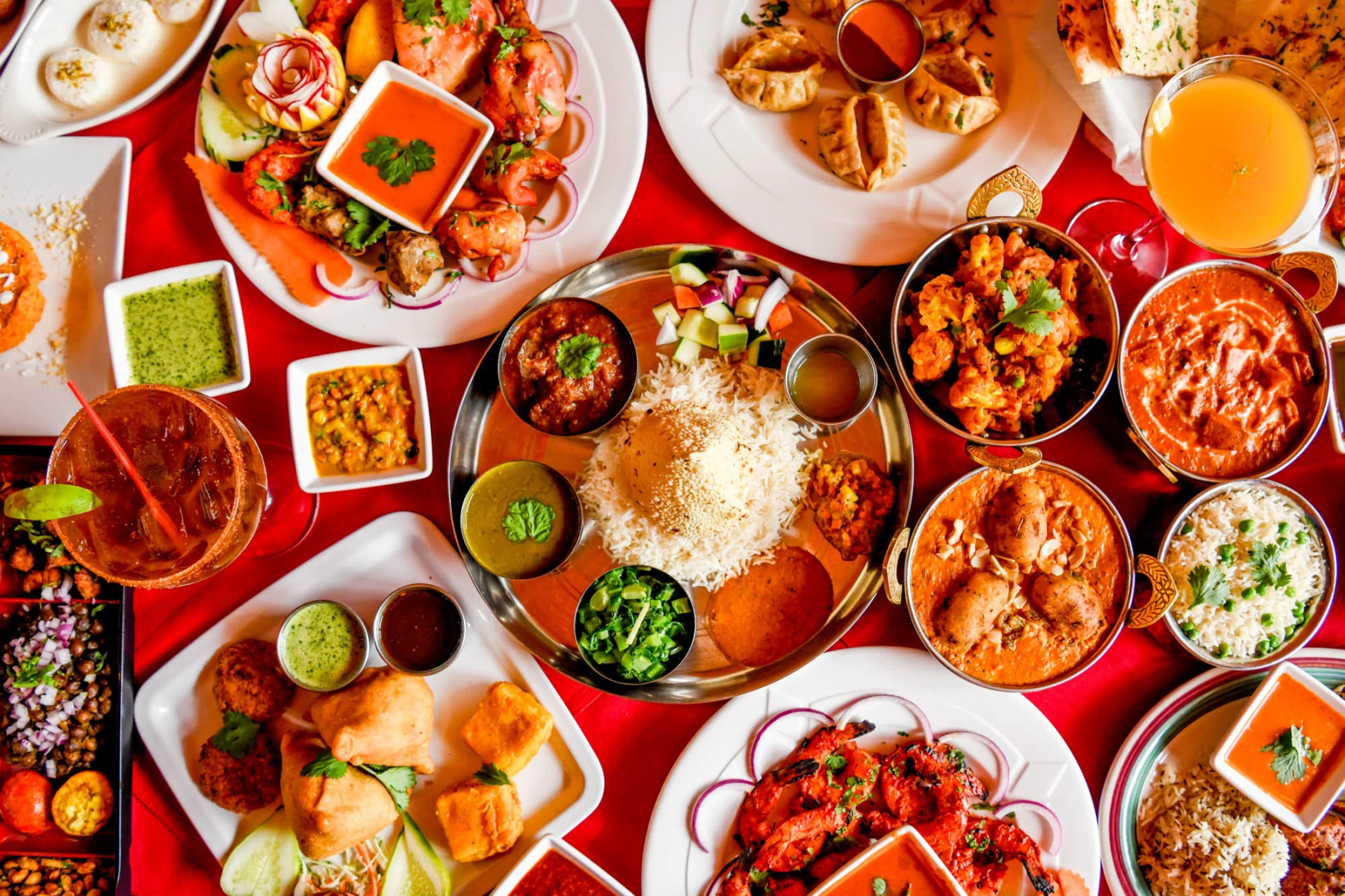 A vibrant spread of Indian dishes including rice, curries, samosas, tandoori shrimp, chutneys, and drinks on a red tablecloth.
