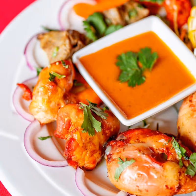 Plate with grilled shrimp garnished with cilantro and red onion slices, served with an orange dipping sauce garnished with cilantro.