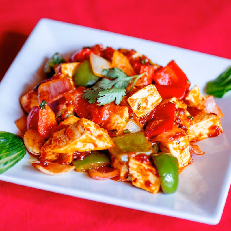 Plate of paneer tikka with red and green bell peppers garnished with cilantro on a white square plate over a red surface.
