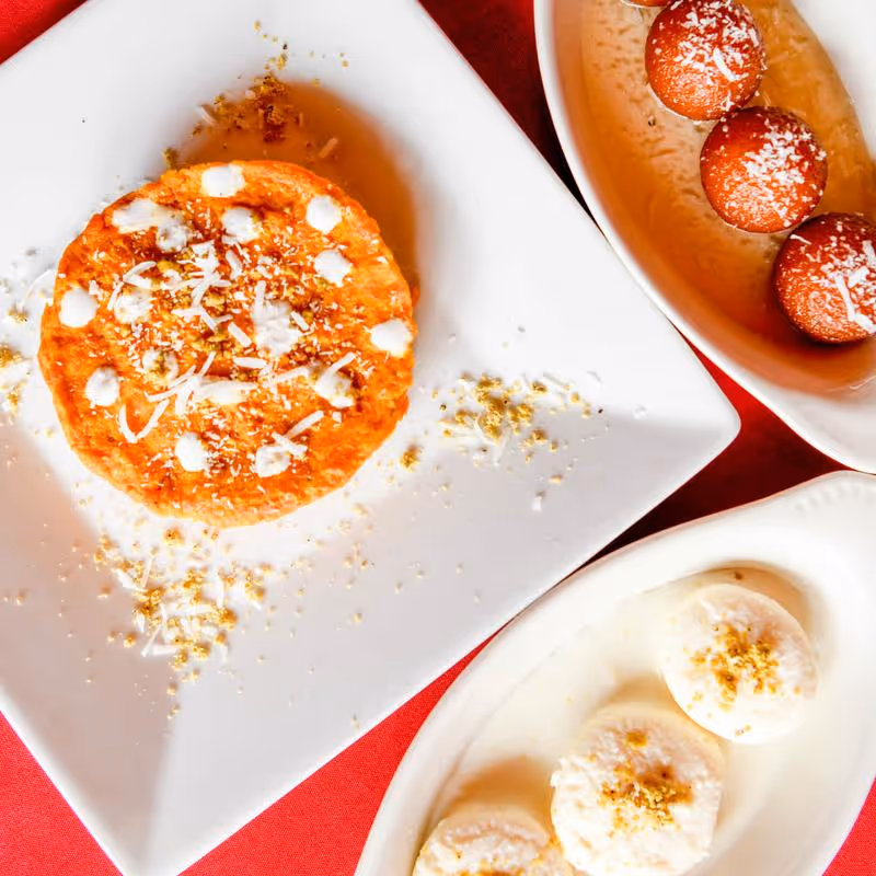 Three Indian sweets: orange-hued dessert topped with white cream on a square plate, round brown fried balls with white topping on an oval plate, and round white sweets with yellow garnish on another oval plate.