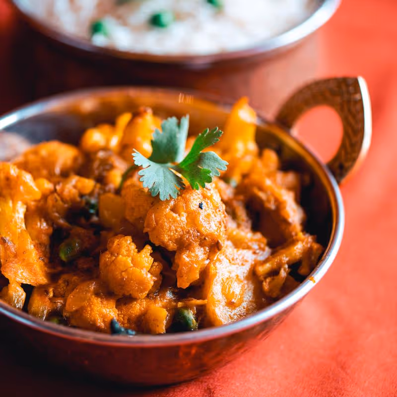 Indian-style cooked cauliflower dish garnished with fresh cilantro in a metal bowl on an orange surface.