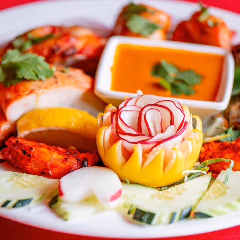 Plate with grilled shrimp, cucumber slices, a carved lemon flower, radish garnish, and a bowl of orange dipping sauce topped with cilantro.