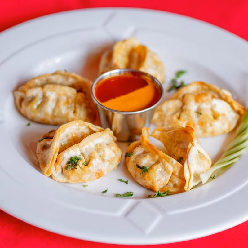 White plate with six fried dumplings garnished with herbs and a small metal cup of red dipping sauce on a red tablecloth.