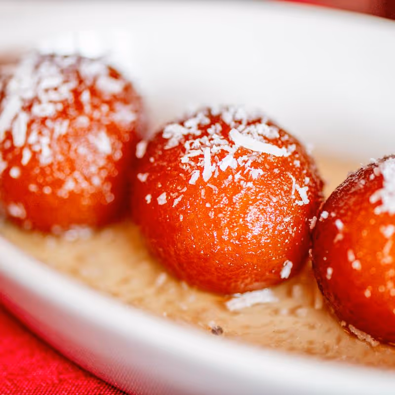 Close-up of three gulab jamuns sprinkled with shredded coconut, served in a white bowl with syrup.