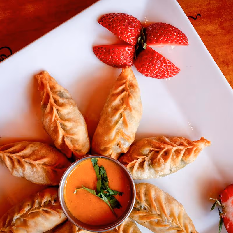 Plate of fried dumplings arranged around a bowl of orange dipping sauce garnished with green herbs, with quartered strawberries on a white plate.