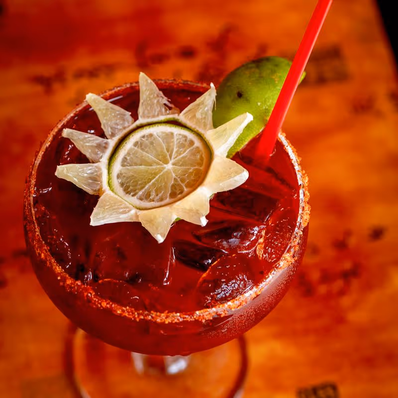 Red cocktail in a glass with a chili powder rim, ice cubes, a red straw, and a lime garnish carved into a sun shape.