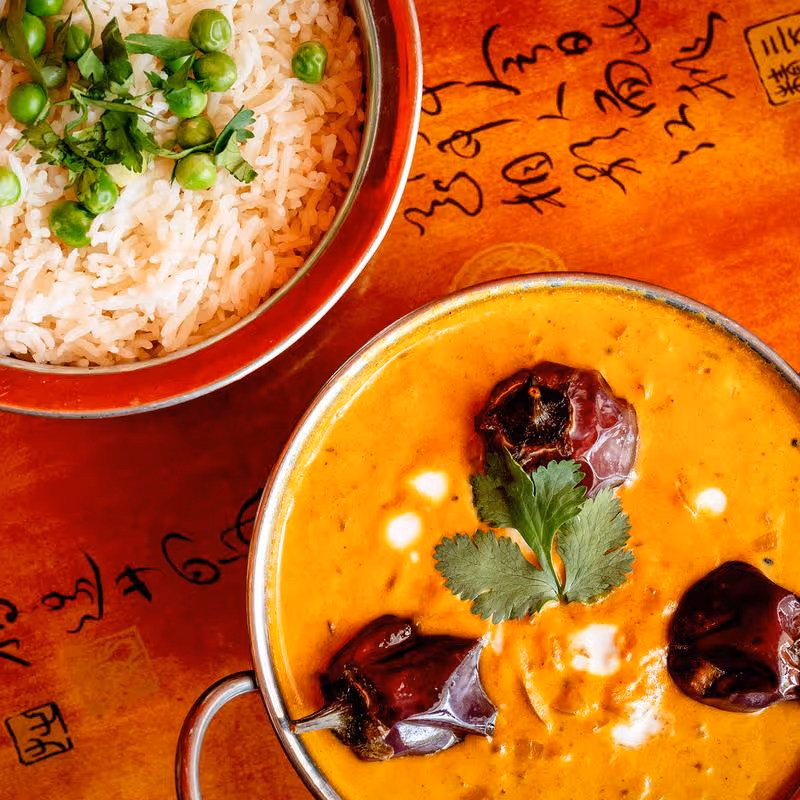 Bowl of yellow curry with red dried peppers and cilantro beside a bowl of white rice topped with green peas and cilantro on a wooden table with calligraphy.