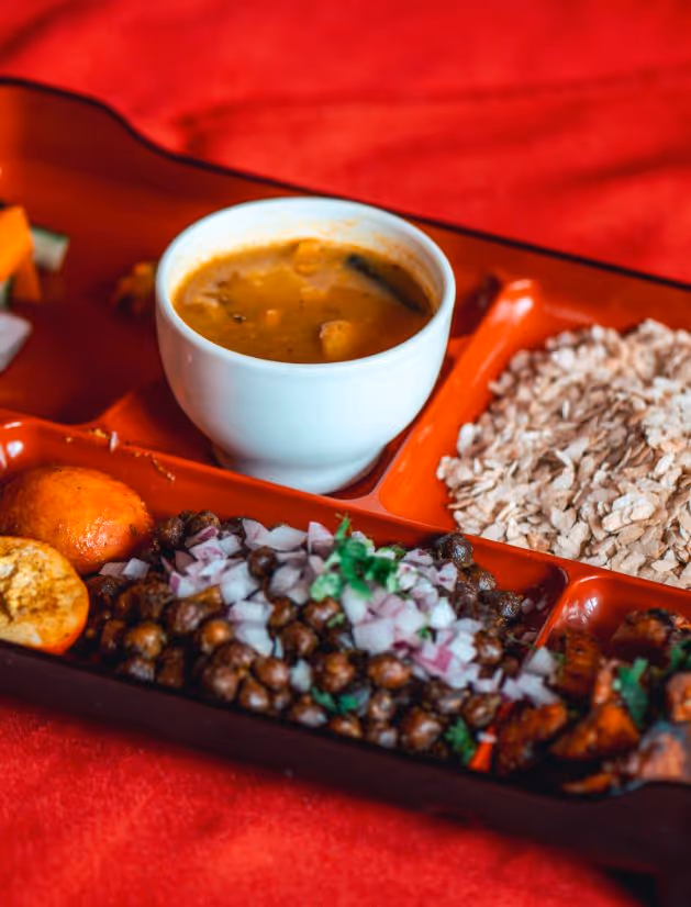 Traditional Indian meal served on a red tray, including a bowl of dal, roasted chickpeas garnished with chopped onions and herbs, a round fried snack, and flaked dry snack.