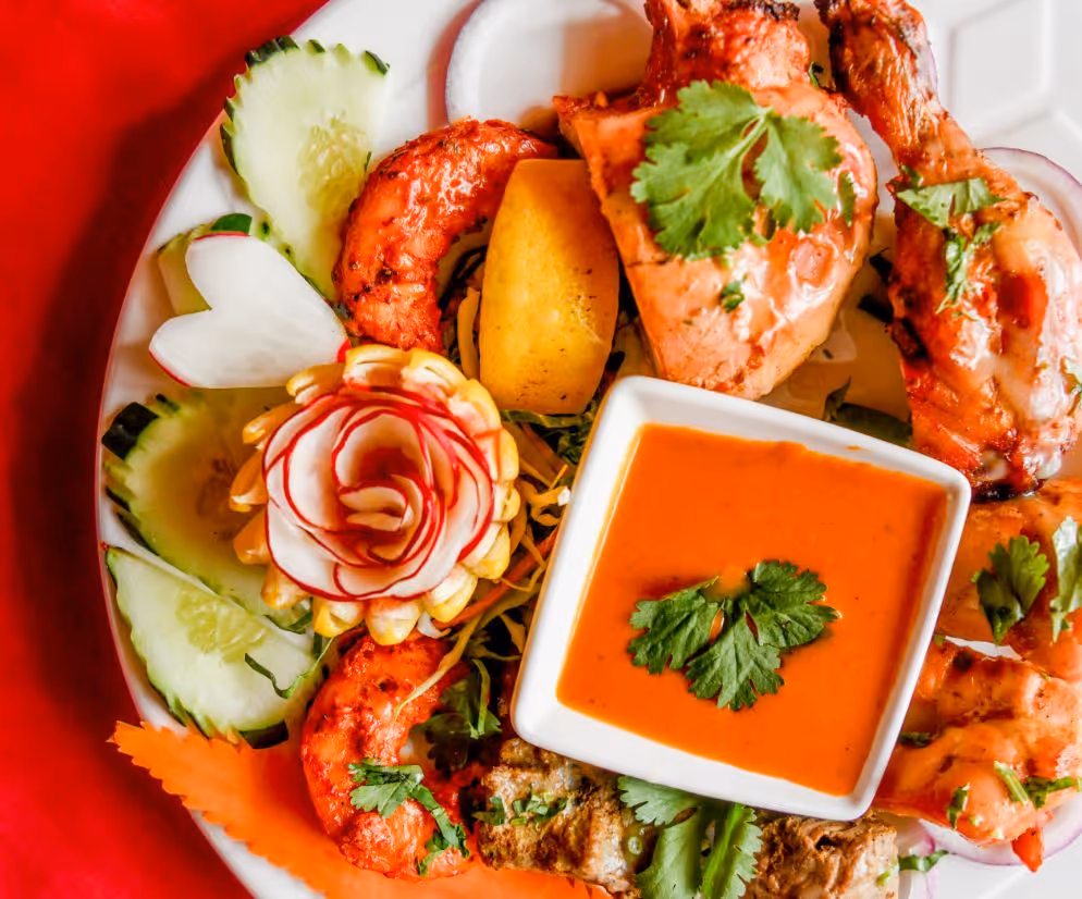 Plate of grilled chicken and shrimp served with cucumber slices, a carved radish rose, carrot garnish, and a bowl of orange dipping sauce garnished with cilantro.