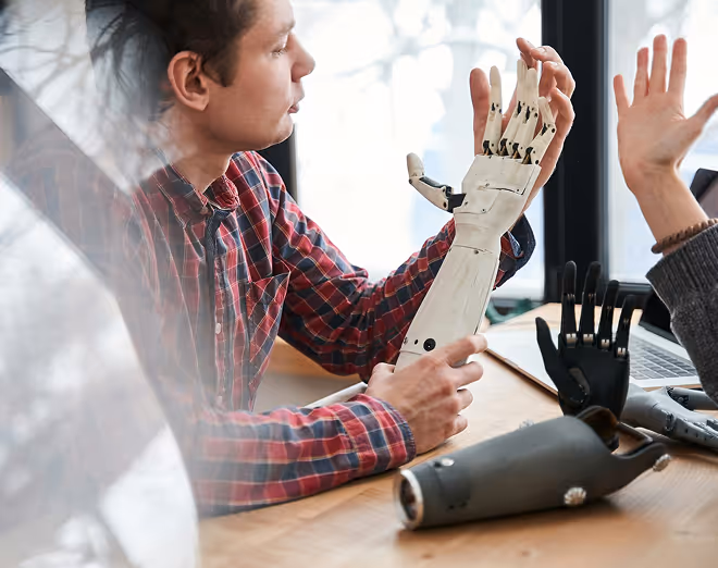Man in plaid shirt examining a white prosthetic arm with a black prosthetic hand on a table.