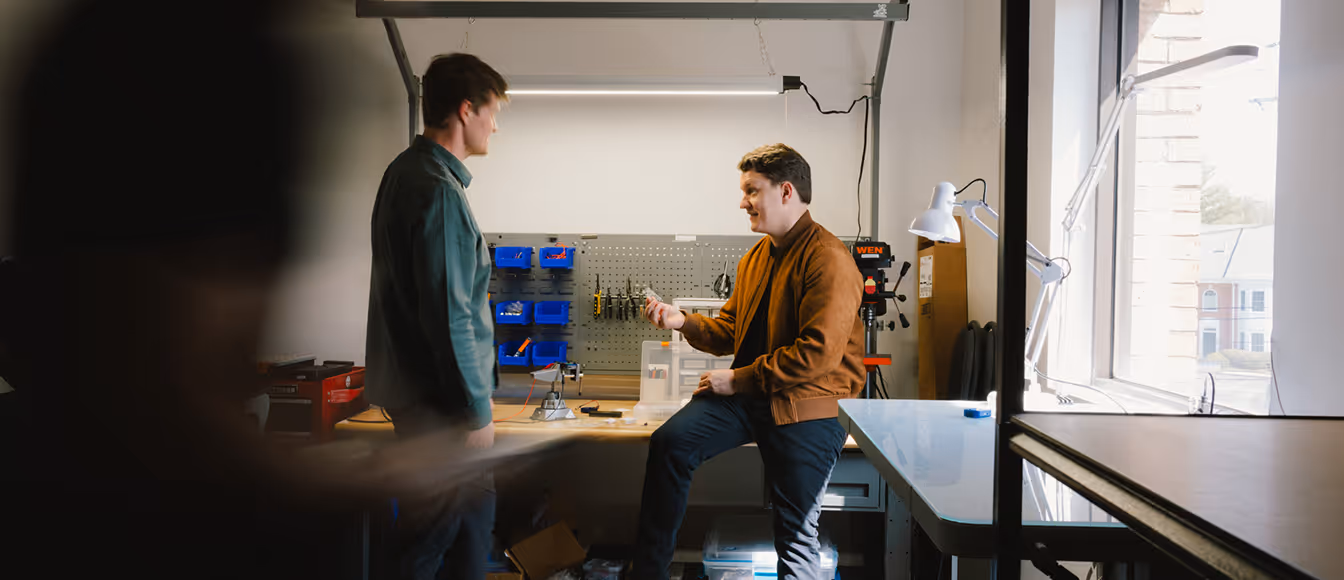 Two men in a workshop talking near a workbench with tools and storage bins on the wall.