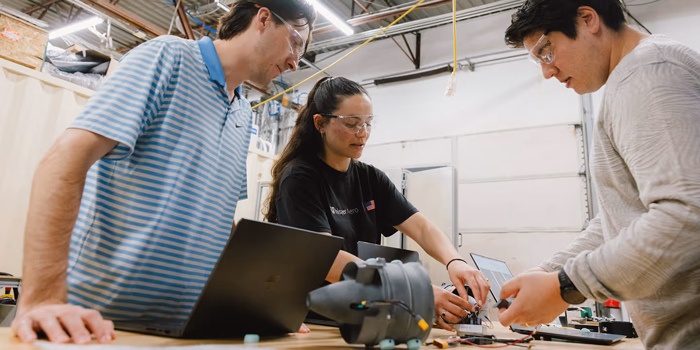Three people wearing safety glasses working on electronic components on a table in a workshop.