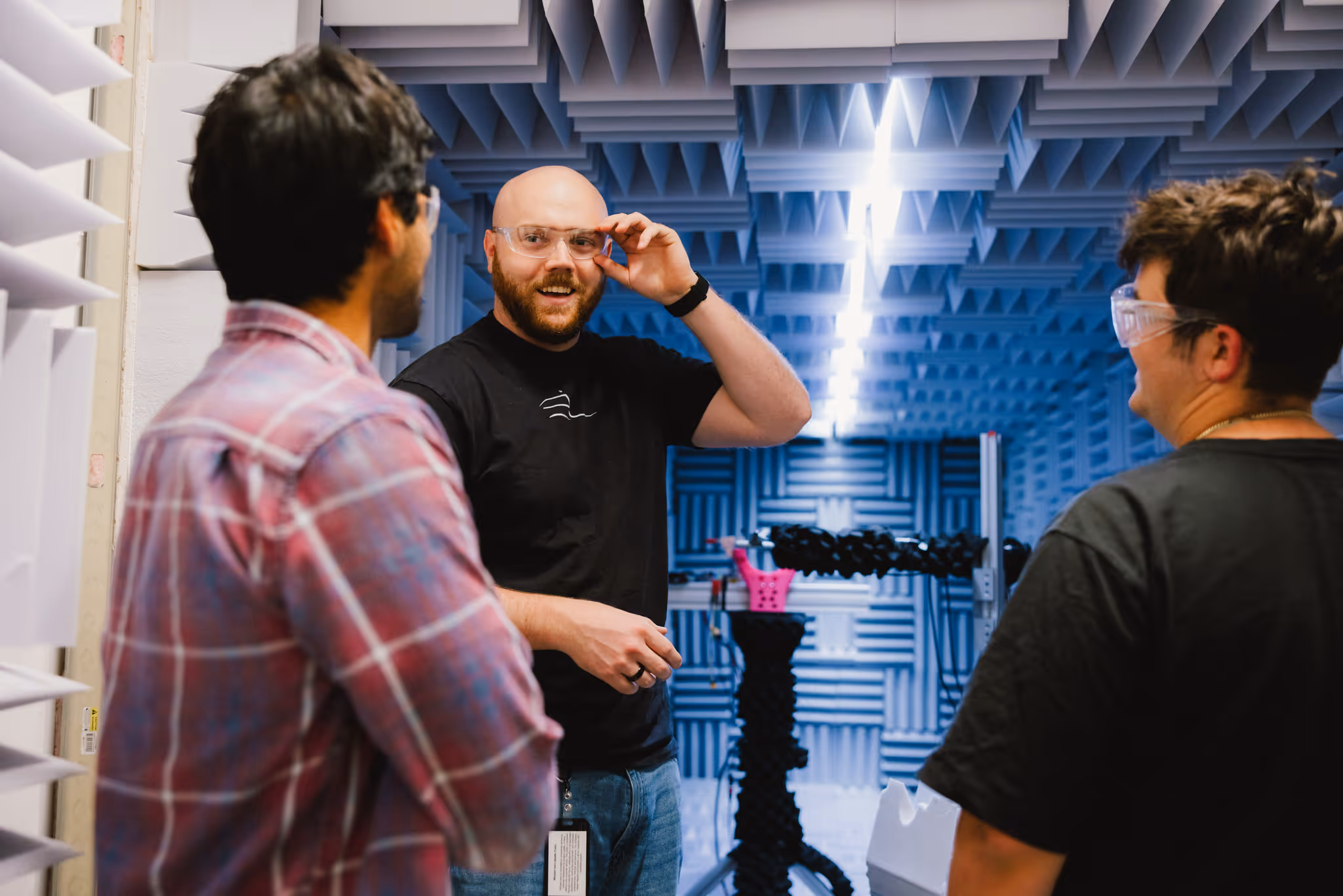 Three people wearing safety glasses talking inside a blue acoustic foam room with equipment.