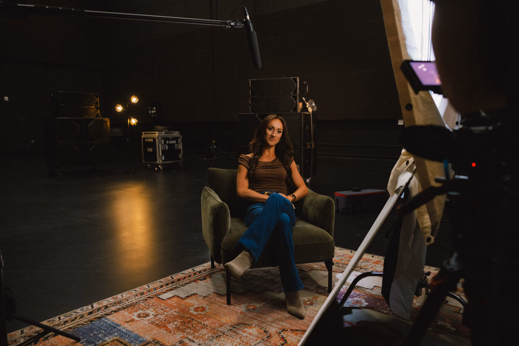Woman sitting in a chair on a patterned rug in a dimly lit studio with filming equipment.