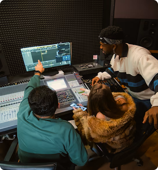 Three people working together in a music studio, focusing on a mixing console and computer screen.