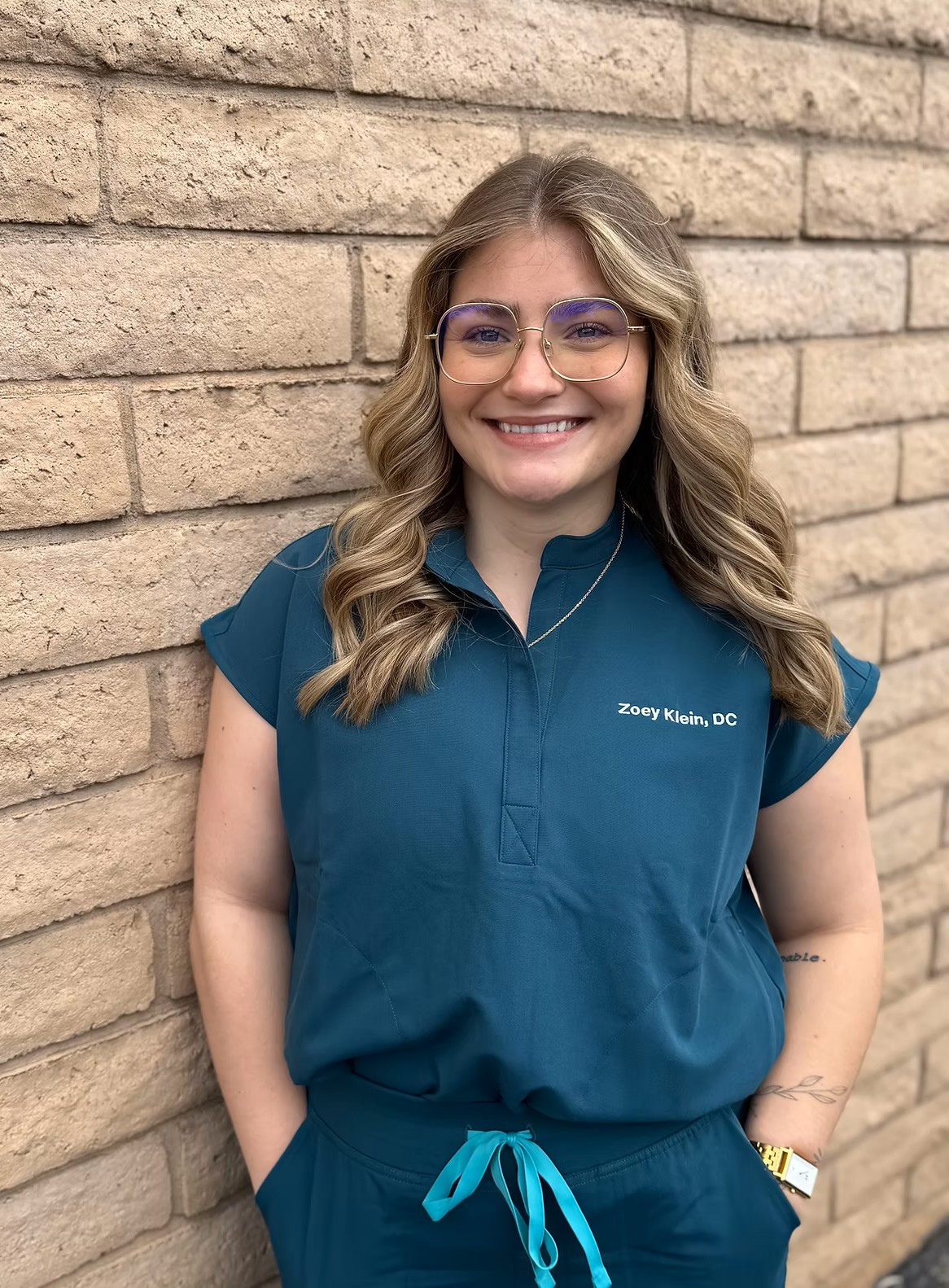 Dr. Zoey, a smiling woman with wavy blonde hair wearing glasses and teal medical scrubs standing against a brick wall.