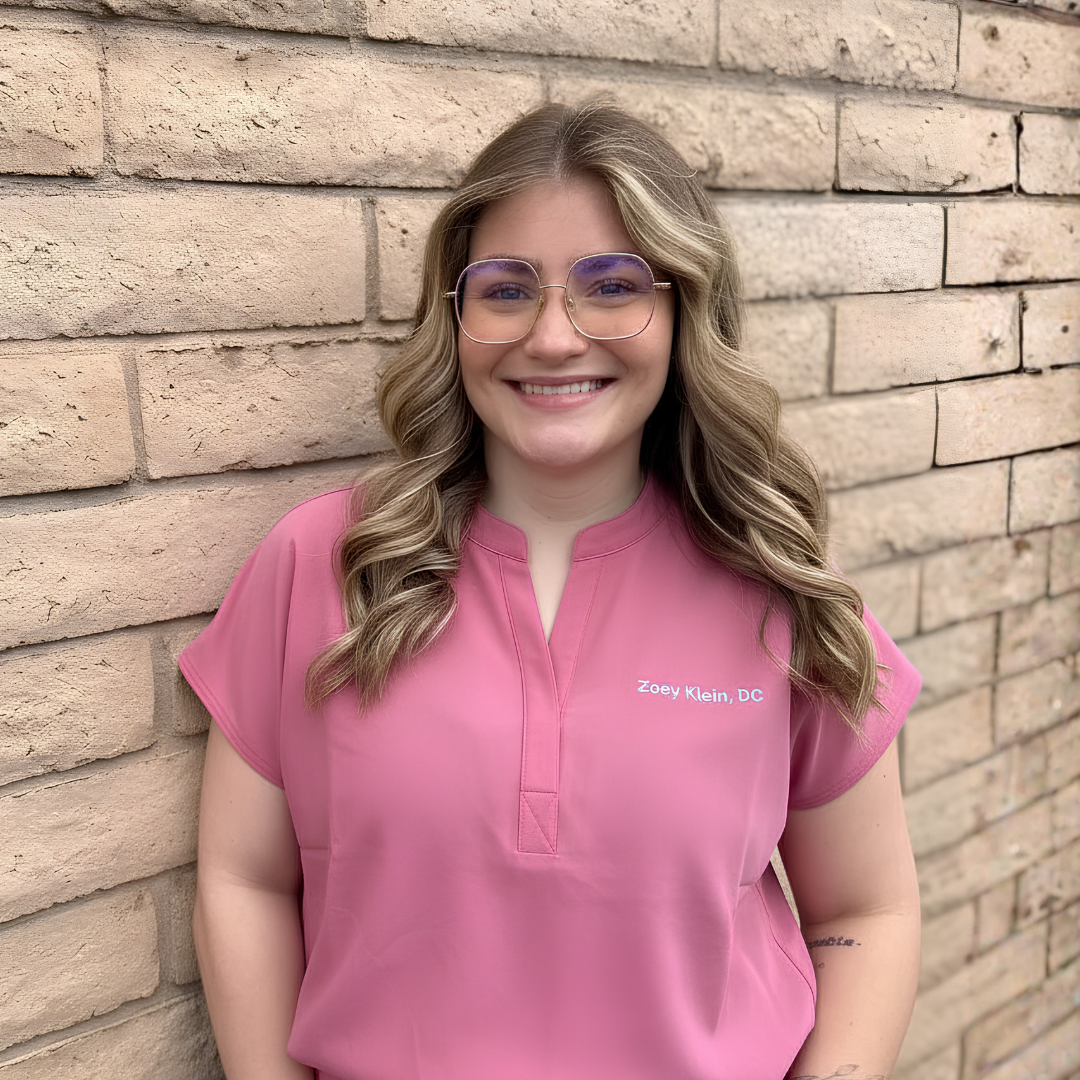 Dr. Zoey, a smiling woman with wavy blonde hair wearing glasses and pink medical scrubs standing against a brick wall.