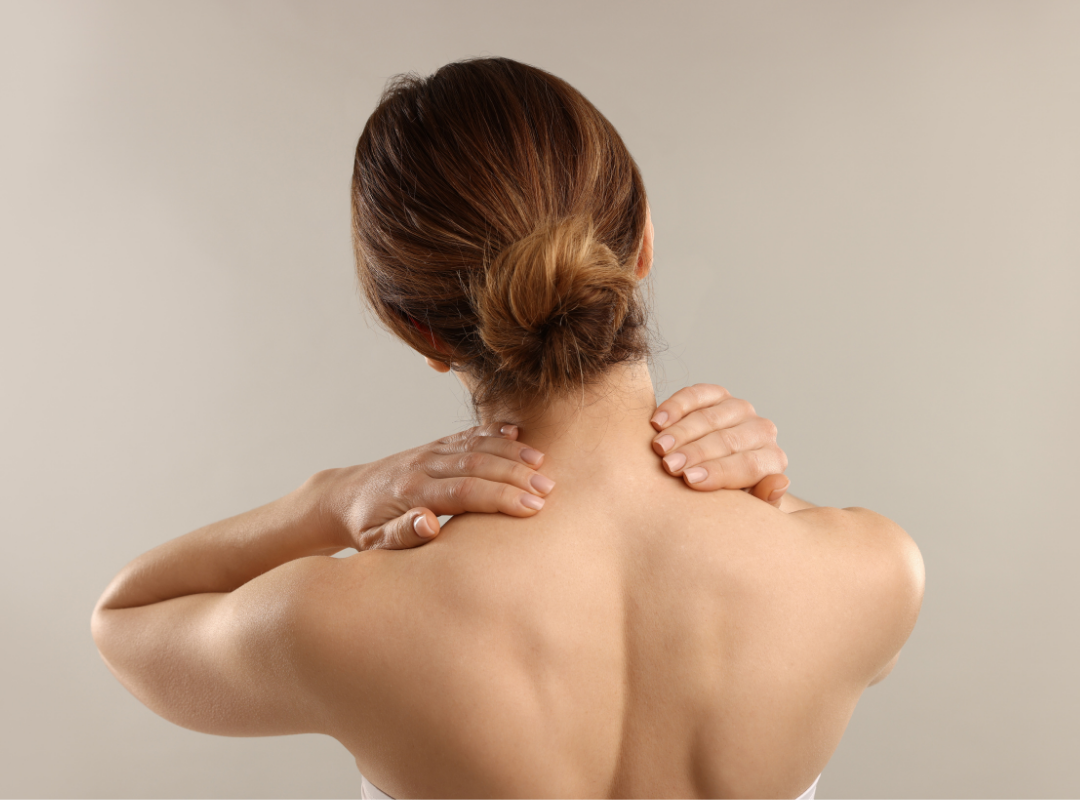 Woman with brown hair in a bun holding her neck and shoulders from the back.