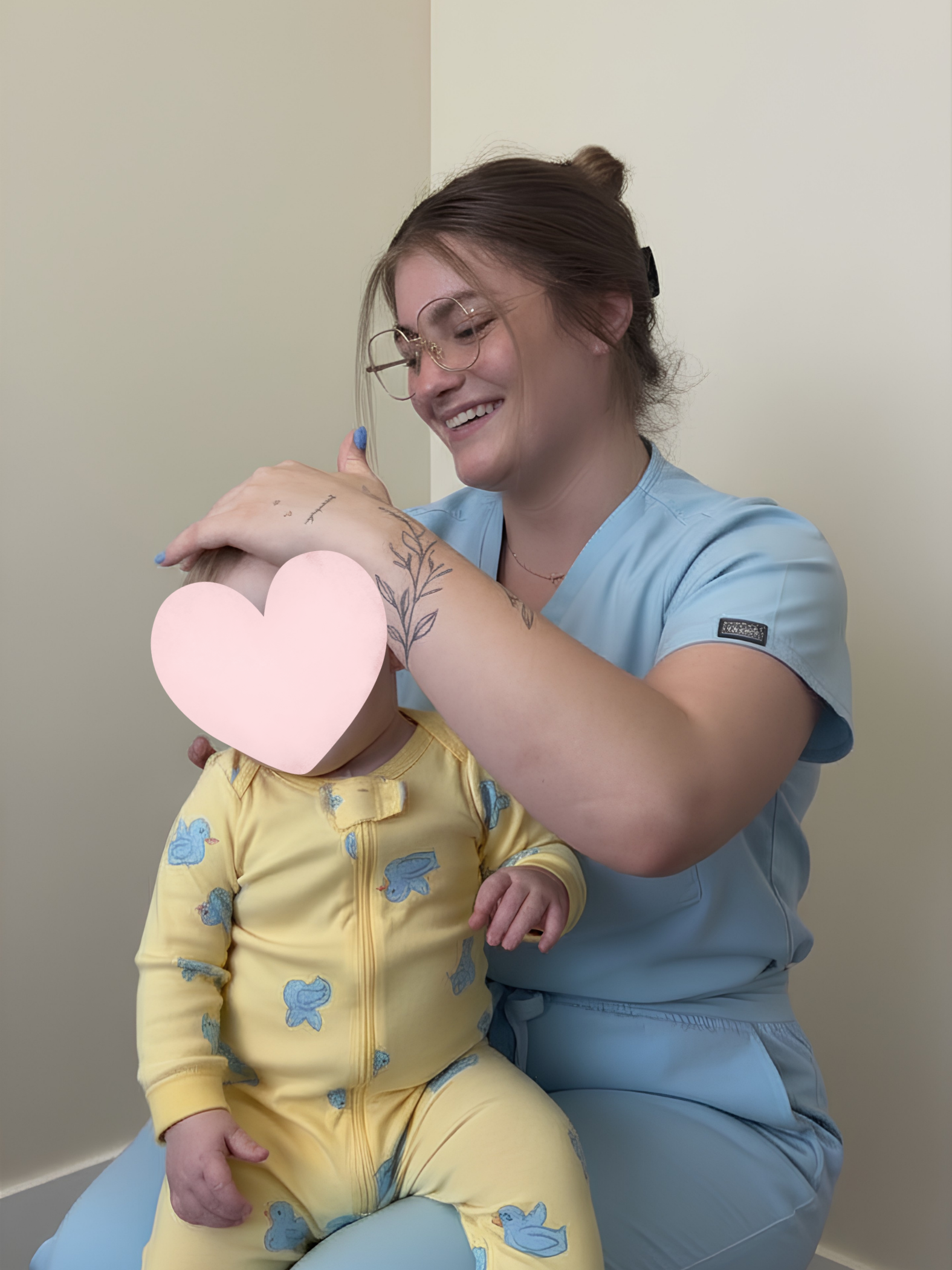 Smiling woman in light blue scrubs gently holding a baby in yellow pajamas with blue duck prints, sitting on her lap.