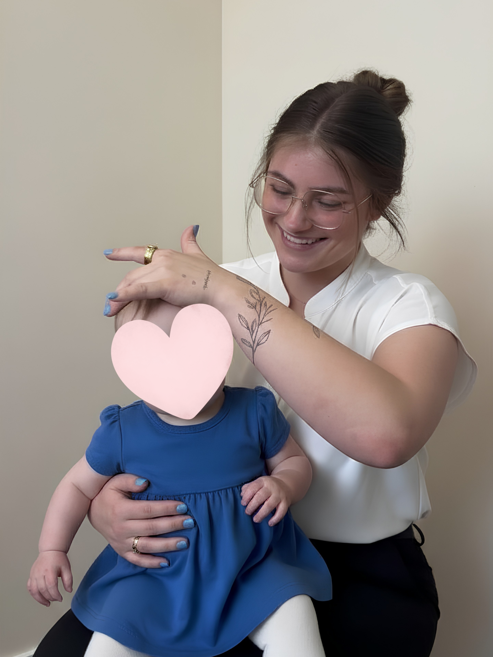 Smiling woman with glasses and tattoos gently holding a baby in a blue dress with a pink heart covering the baby's face.