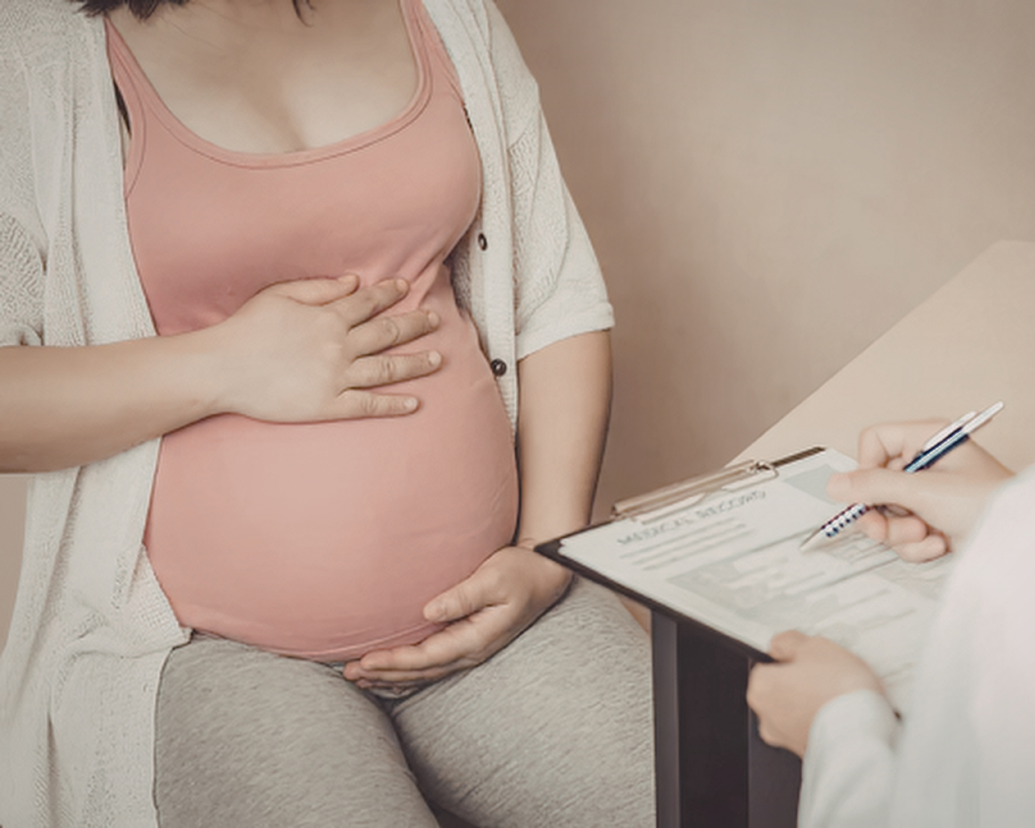 Pregnant woman holding her belly during a medical consultation with a healthcare professional writing on a clipboard.