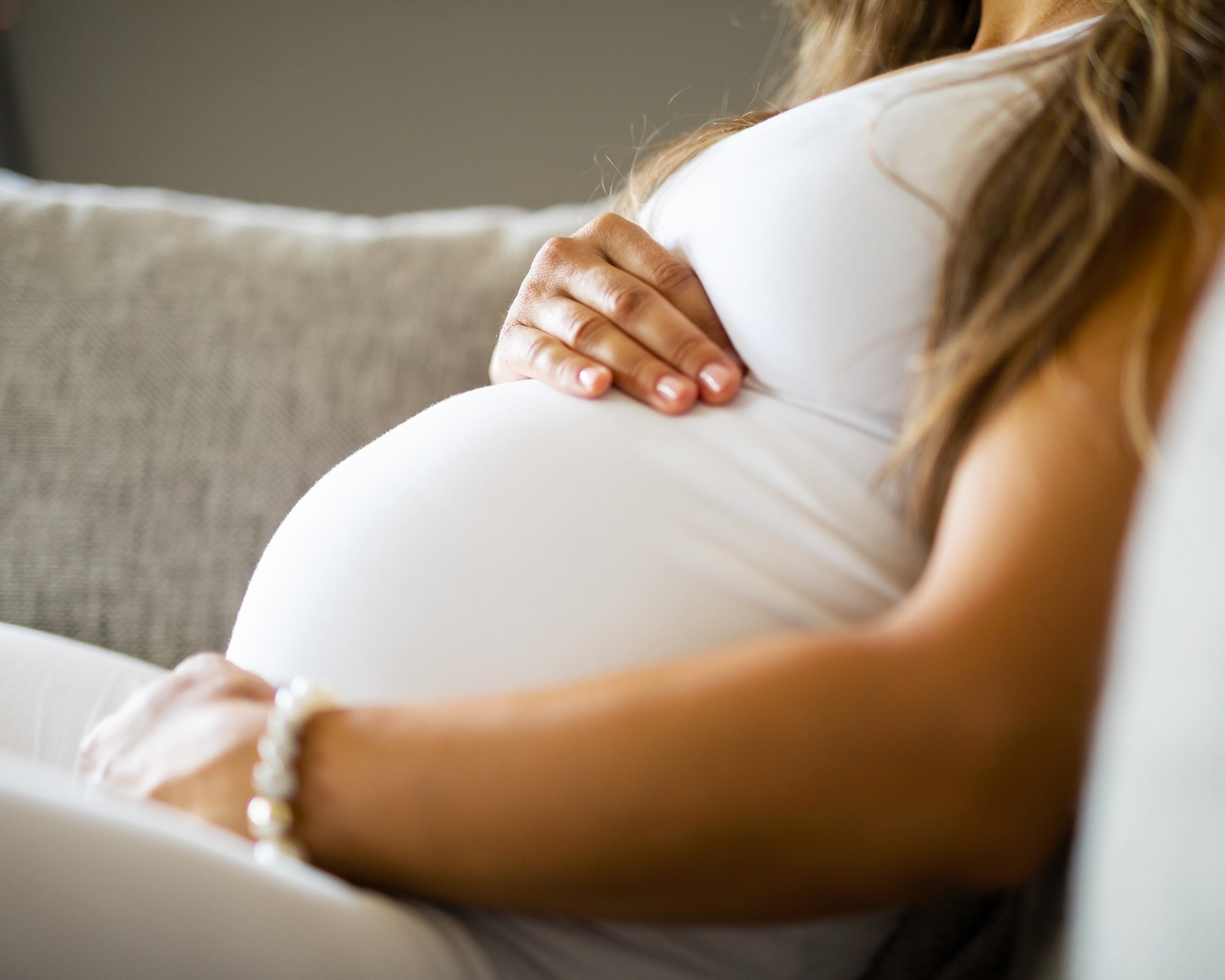 Close-up of a pregnant woman in a white dress sitting on a couch, gently resting her hand on her belly.