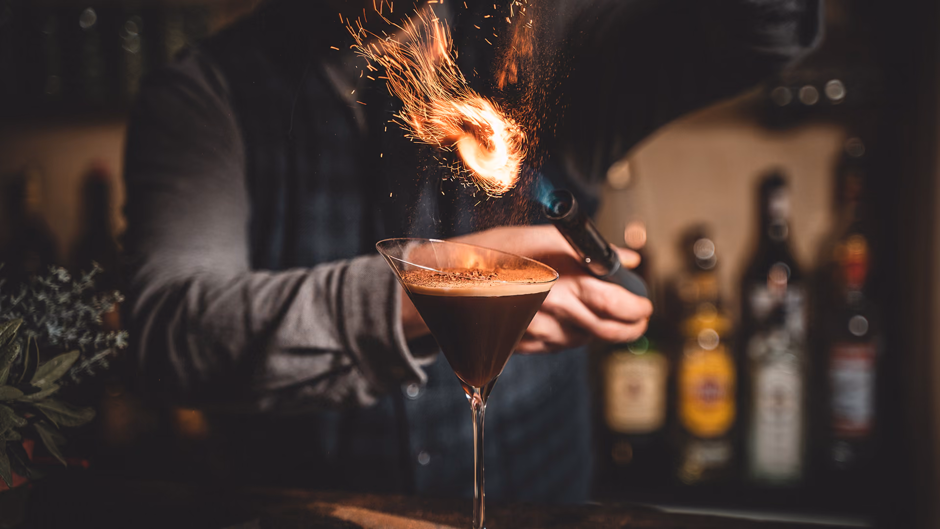 Ein Barkeeper flambiert eine Orangenschale über einem Martiniglas und lässt Funken über der dunklen Cocktailbar aufsteigen; die Szene wirkt dramatisch und kultiviert.