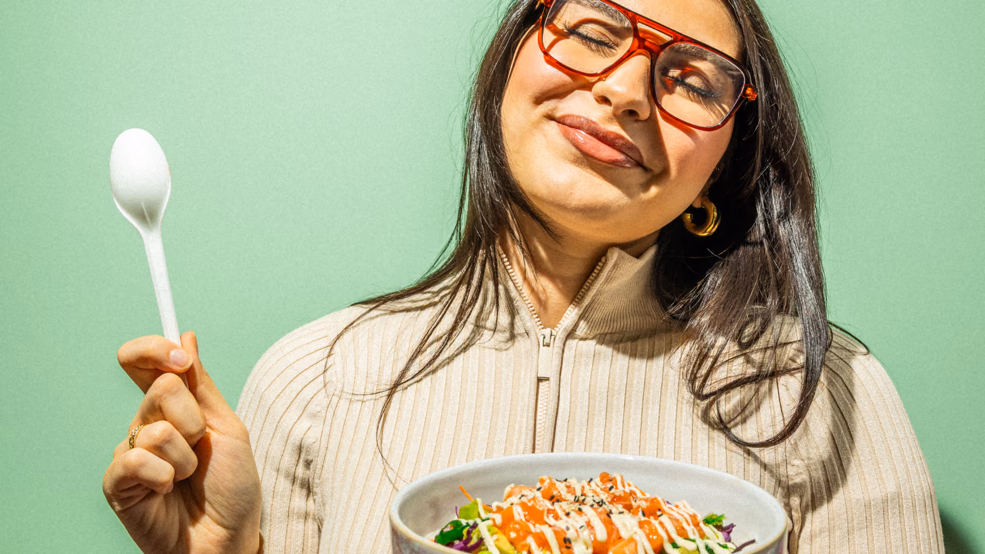 Eine lächelnde Frau mit roter Brille hält vor einem mintgrünen Hintergrund einen Löffel und eine Schale mit buntem Essen in der Hand, was eine fröhliche, einladende Stimmung schafft.