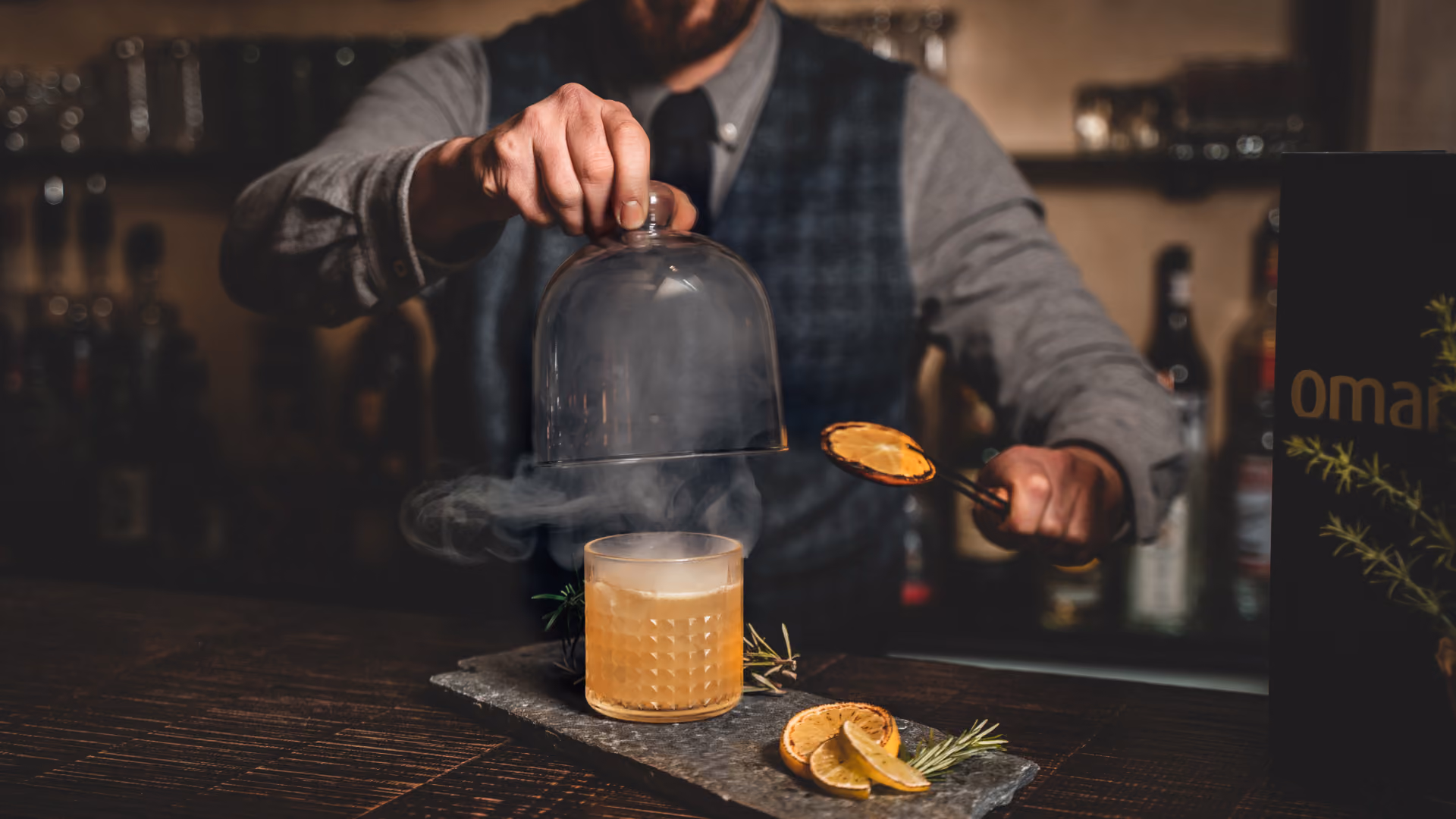 Ein Barkeeper hebt eine Glasglocke von einem bernsteinfarbenen Cocktail und hält dabei eine Orangenschale in der Hand; Zitronenscheiben und Rosmarin schmücken die dunkle, stimmungsvolle Bar.