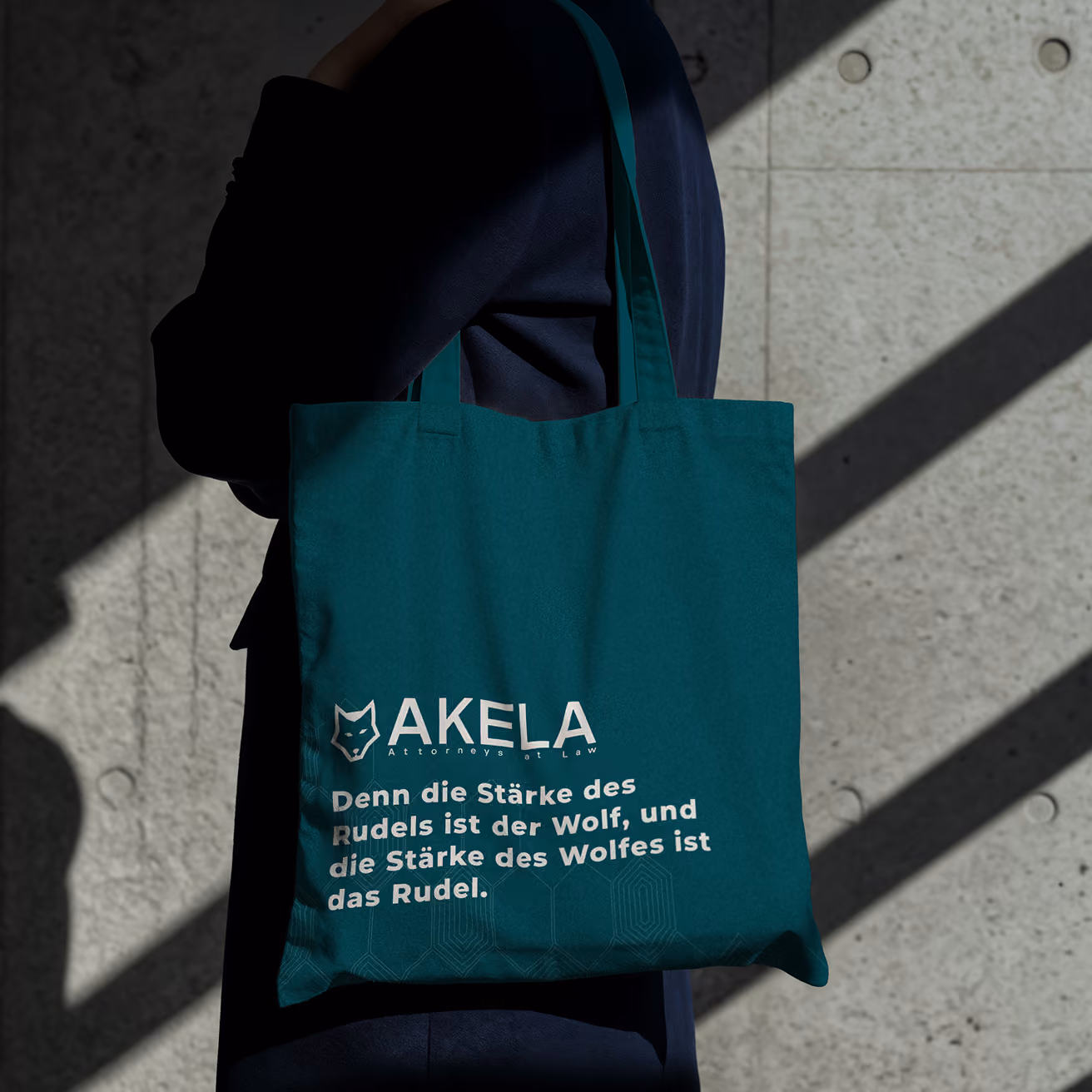 Petrolfarbene AKELA-Tragetasche mit deutschem „Wolfsrudel“-Slogan, getragen von einer im Schatten befindlichen Person vor einer Betonwand mit markanten diagonalen Schatten.