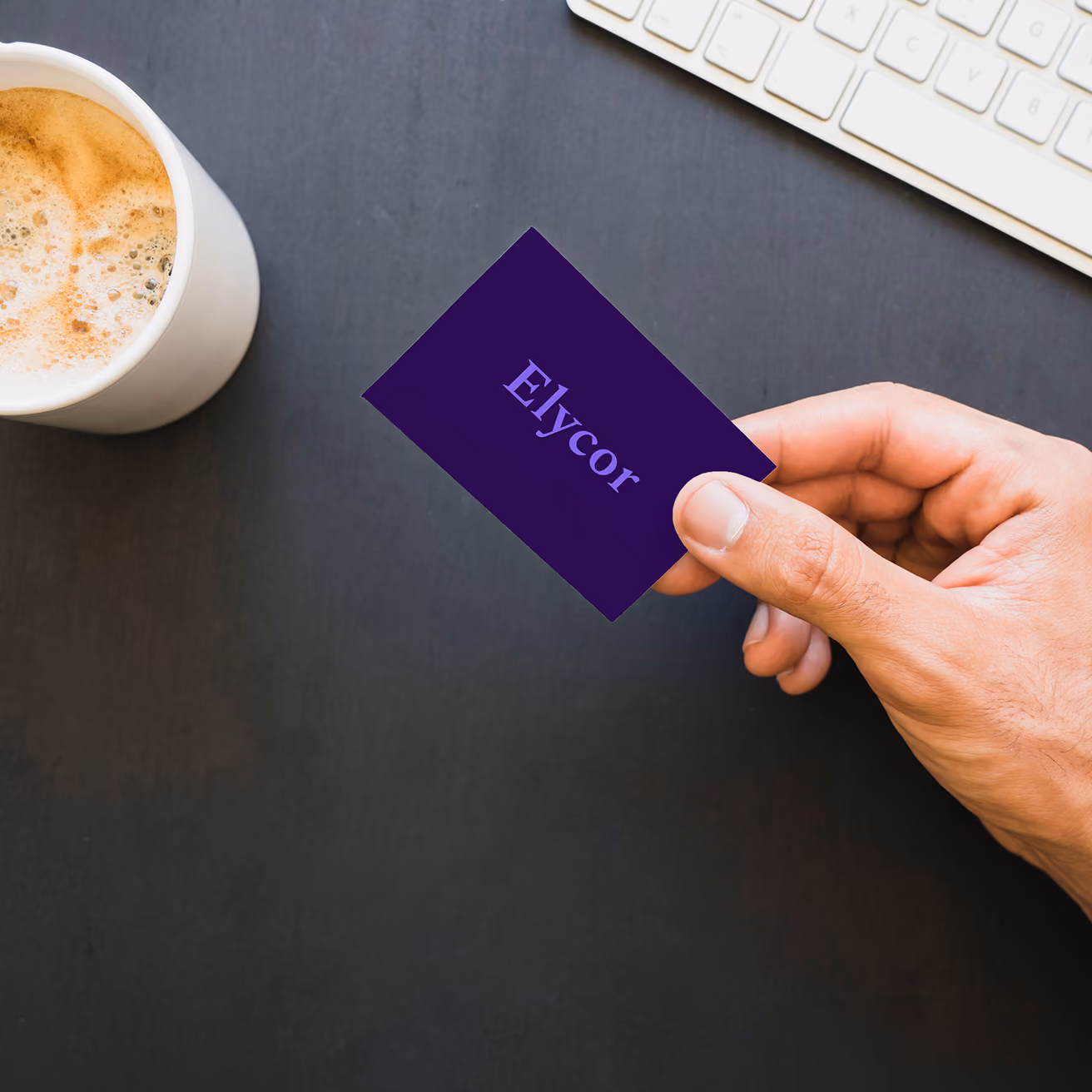 Eine Hand hält eine violette „Elycor“-Visitenkarte über einen dunklen Schreibtisch; Kaffee und Tastatur befinden sich in der Nähe, was eine professionelle Büroumgebung vermittelt.