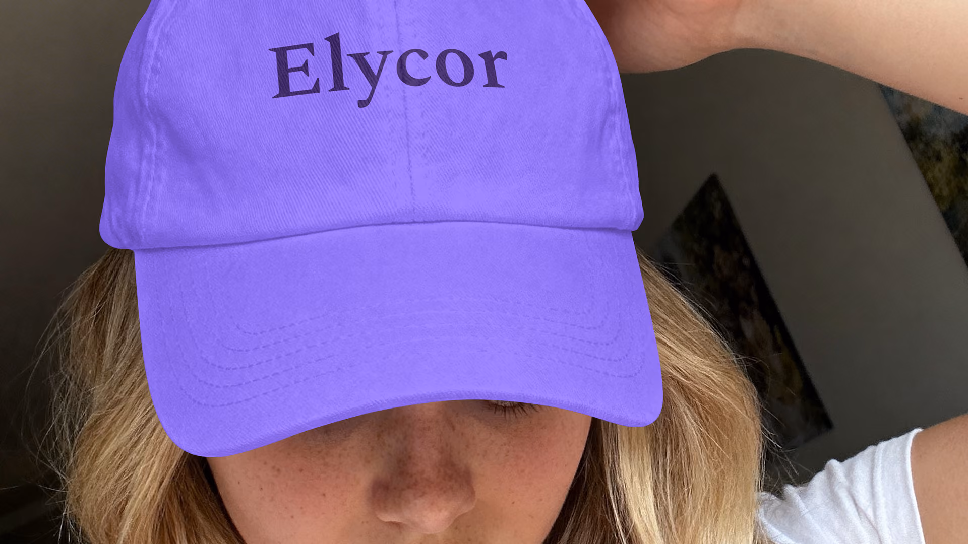 Nahaufnahme einer Person mit blondem Haar und weißem Hemd, die eine leuchtend violette Baseballkappe mit der Aufschrift „Elycor“ trägt; die Aufnahme entstand in einem schwach beleuchteten Innenraum.
