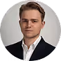 Professionelles Porträtfoto eines jungen Mannes mit neutralem, ernstem Ausdruck, bekleidet mit einem dunklen Anzug und einem weißen Hemd, vor hellgrauem Hintergrund.