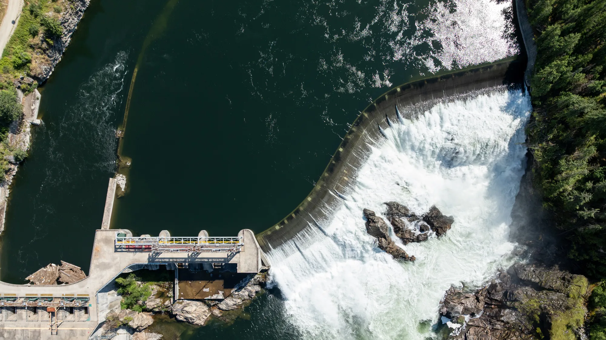 Aerial view of a curved dam releasing water into a river surrounded by trees and rocks.