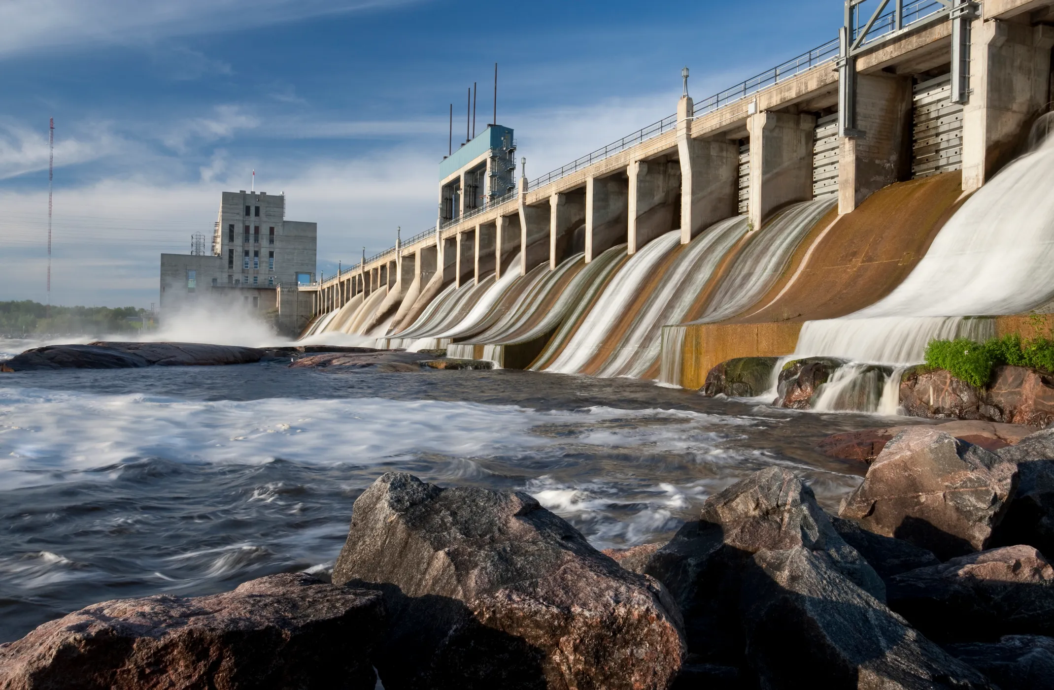 Water flows over a large concrete dam with a building and rocks in the foreground under blue sky.