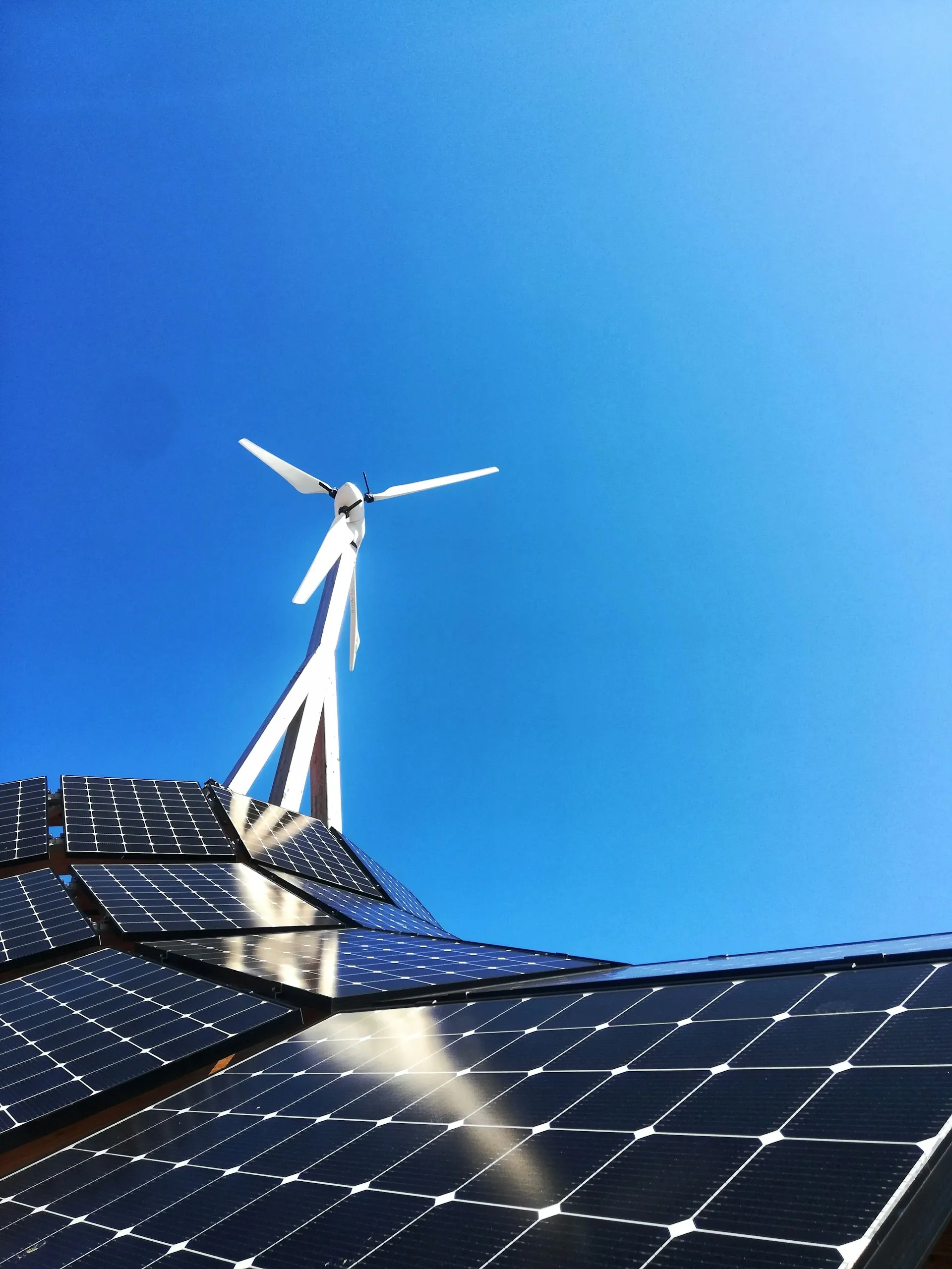 Solar panels reflect sunlight beneath a white wind turbine under a clear blue sky.