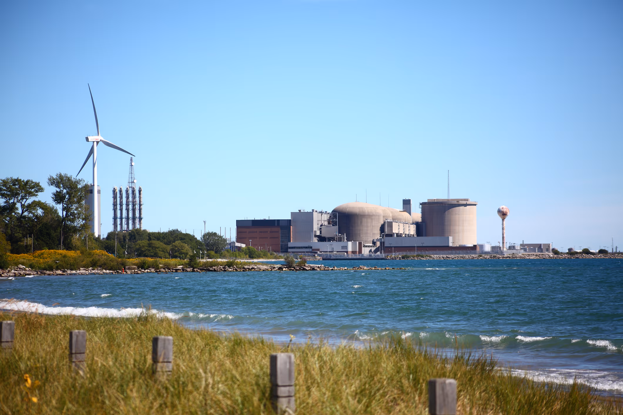Waterfront view of a nuclear power plant with storage tanks and a wind turbine under a clear sky.