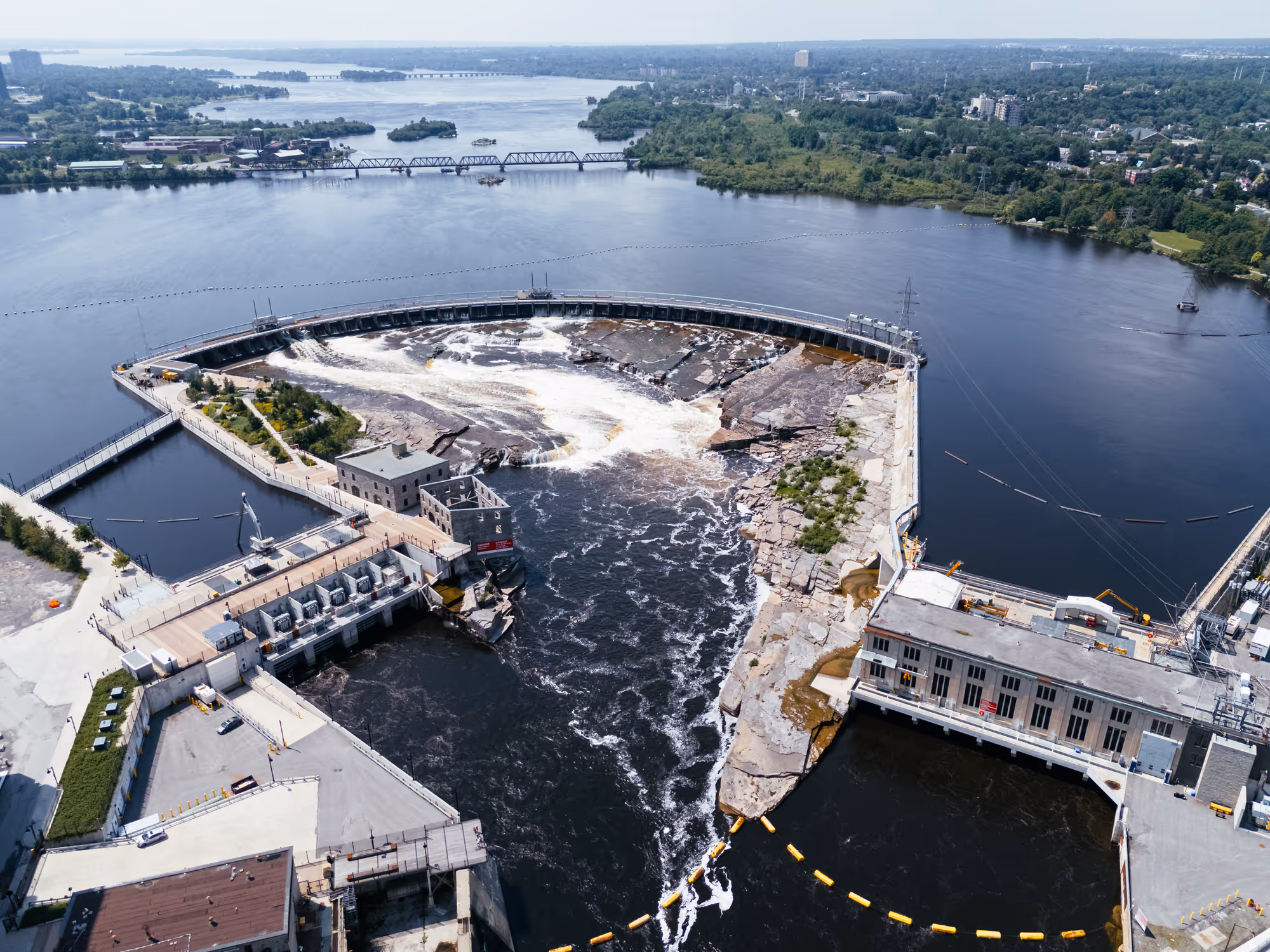 Aerial view of a curved hydroelectric dam with water flowing and surrounding buildings and greenery.