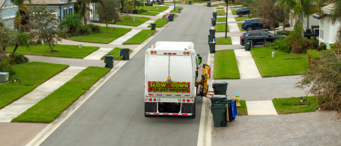 Garbage truck collecting bins on a residential street lined with houses and green lawns.