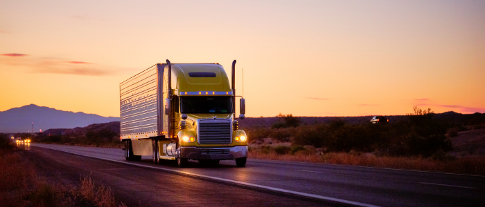 Yellow semi truck driving on highway at sunset with mountains in the background.