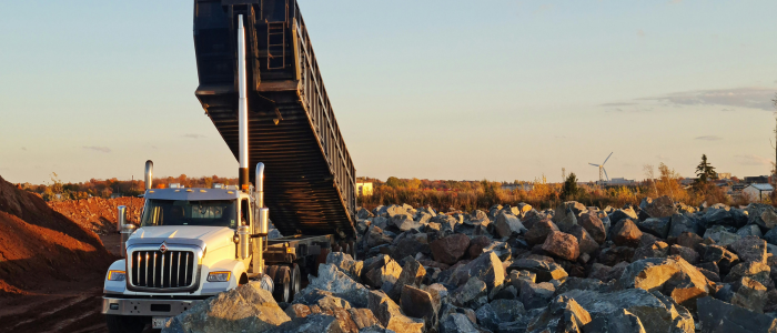 White dump truck unloading rocks on a construction site during sunset with wind turbines in the background.