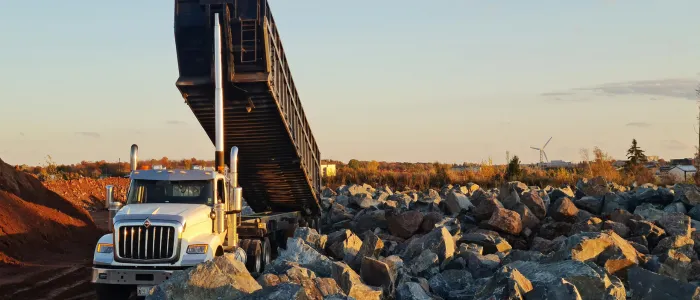 White dump truck unloading rocks on a construction site during sunset with wind turbines in the background.