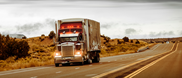 Large semi-truck with red lights driving on a two-lane highway through a dry, hilly landscape.